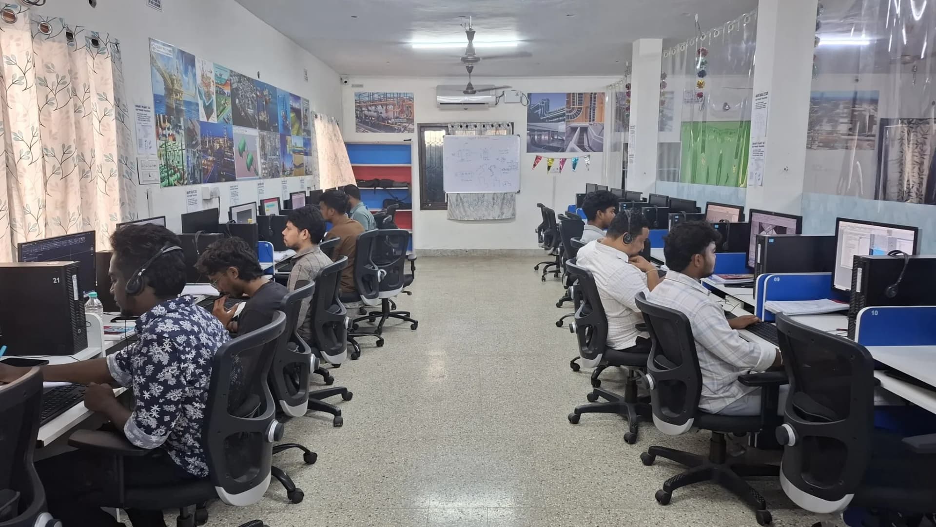 Students wearing headphones sit at rows of desks using desktop computers in a computer lab with posters on the walls behind them.
